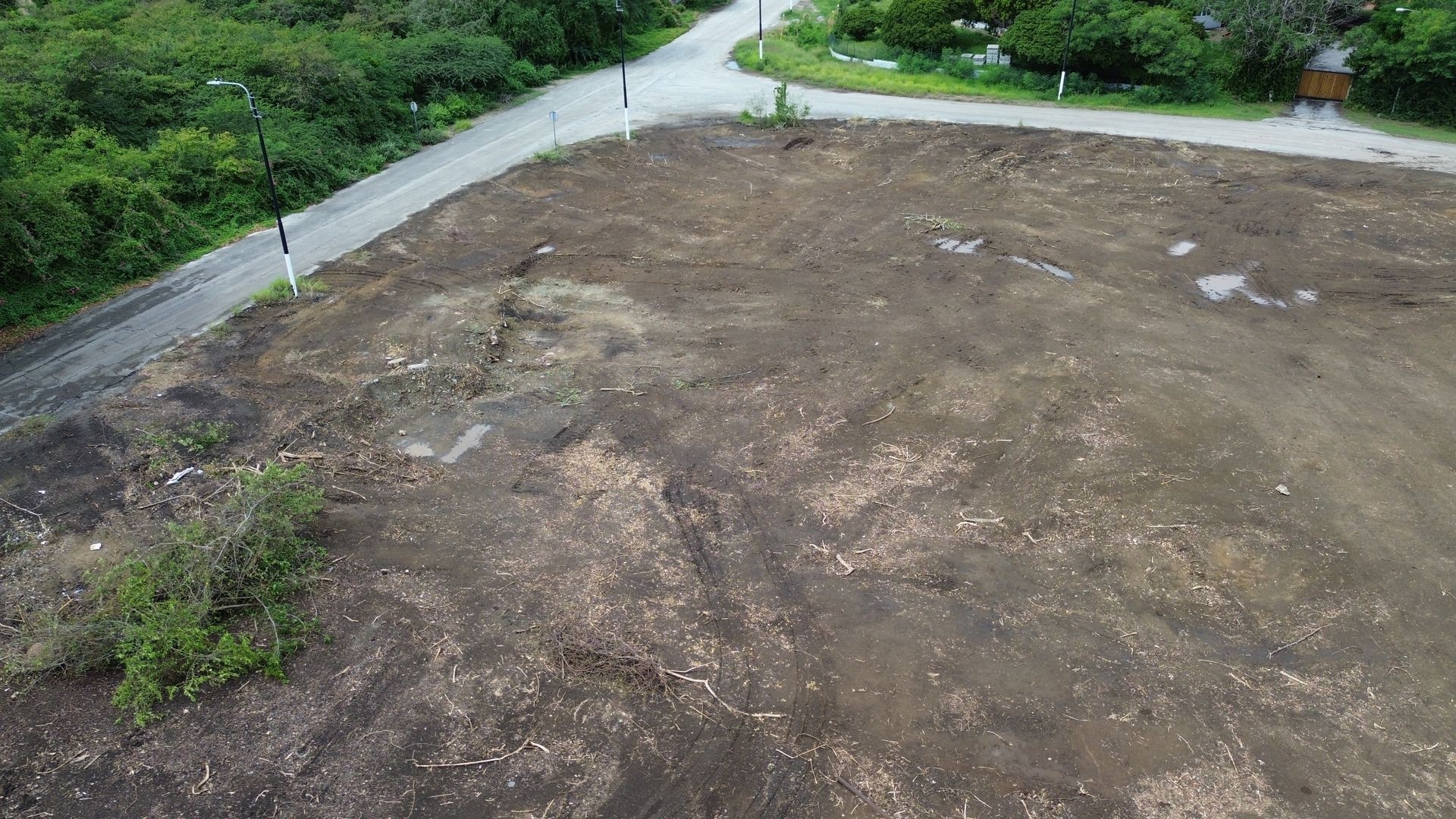 Aerial view of an empty dirt lot with surrounding greenery and a road adjacent to the lot.