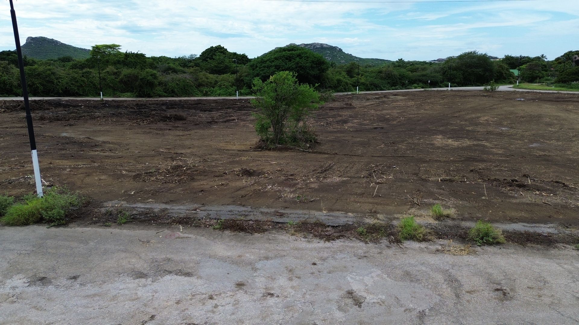 Cleared land with sparse vegetation surrounded by green trees and distant hills under a cloudy sky.